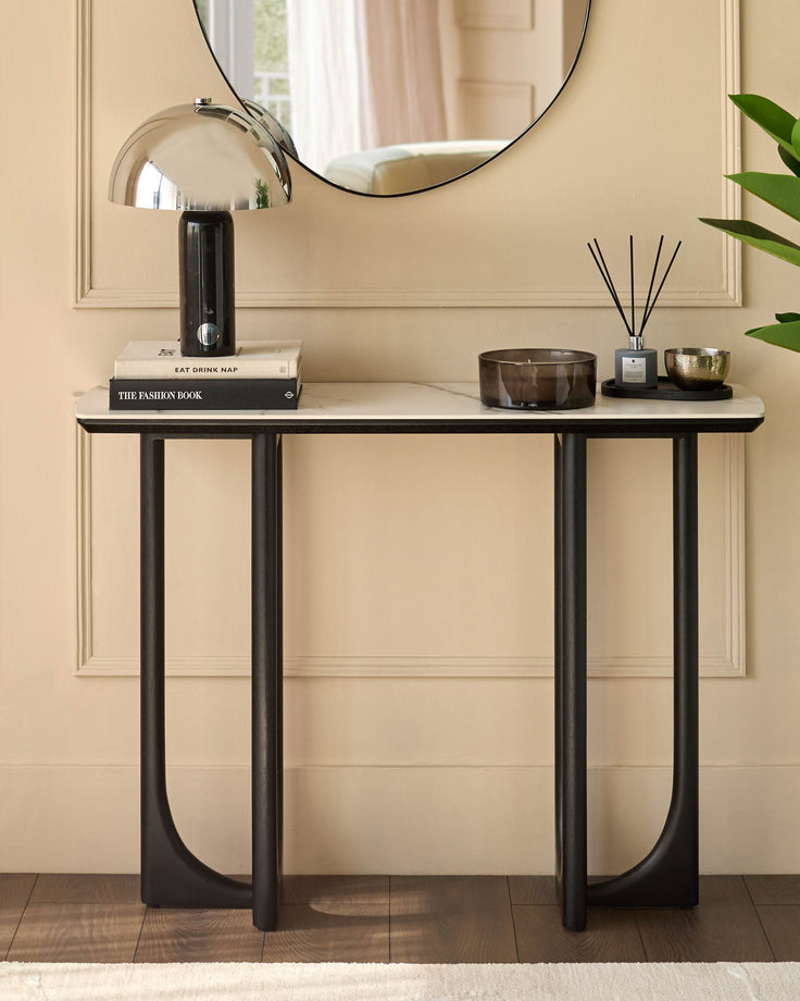 A console table with marble top and black legs displaying books, a lamp and fragrance diffusers.