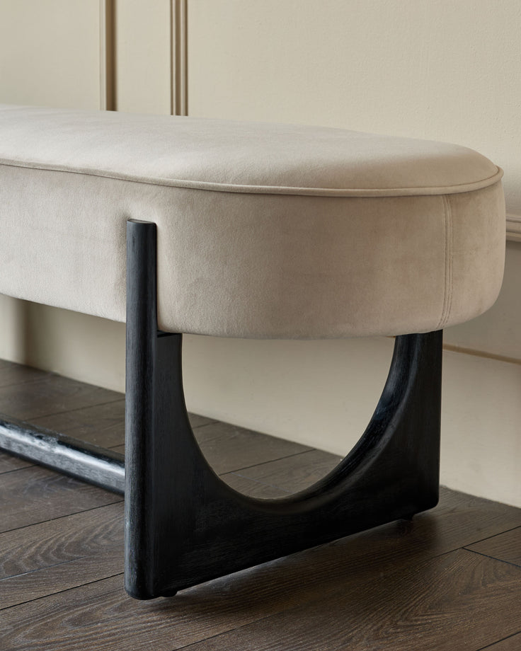 Close-up of a beige upholstered bench with curved black legs on a dark wooden floor.
