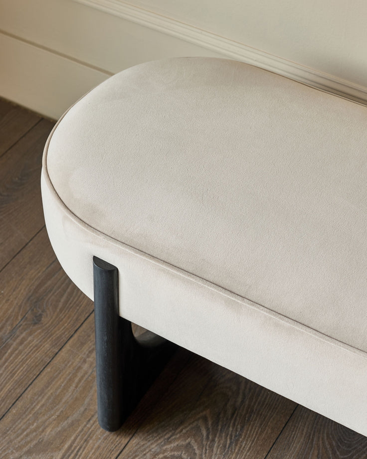 A stylish, beige-toned velvet bench with dark wood legs sits on a wooden floor.