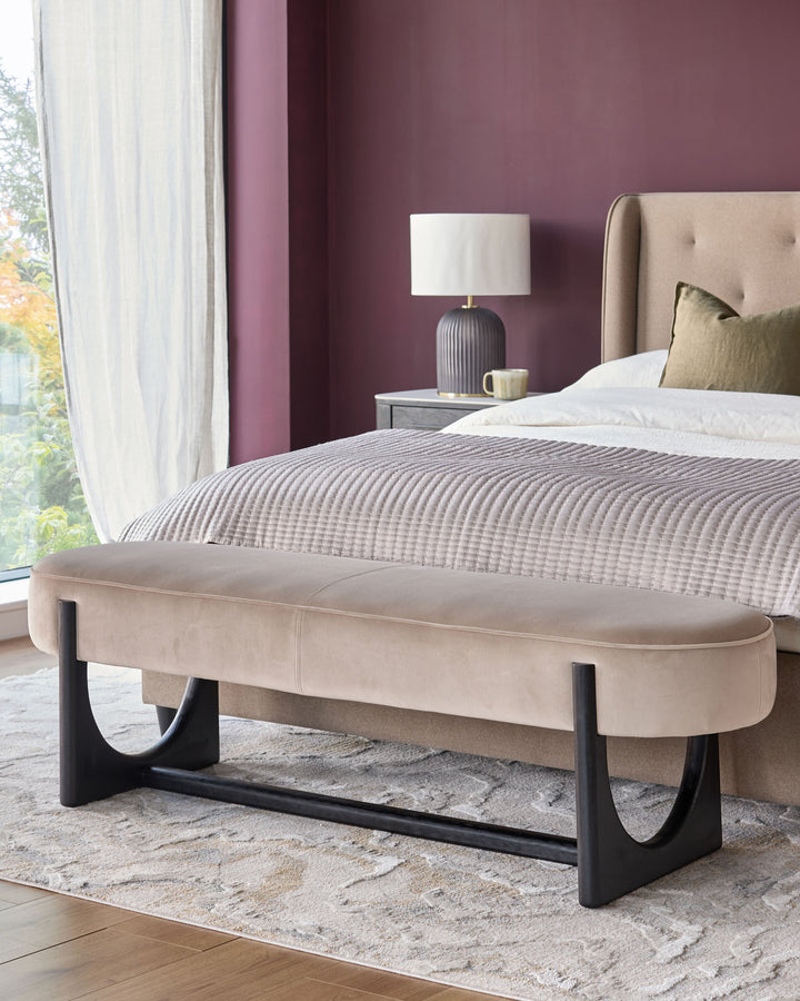 Bedroom with upholstered bed and bench, side table with lamp. Window with draped sheer curtains.