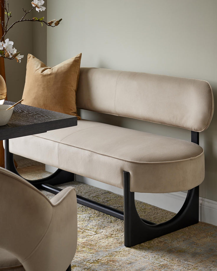 A stylish off-white upholstered bench with black metal legs sits on a patterned rug. An armchair and dark table are nearby.
