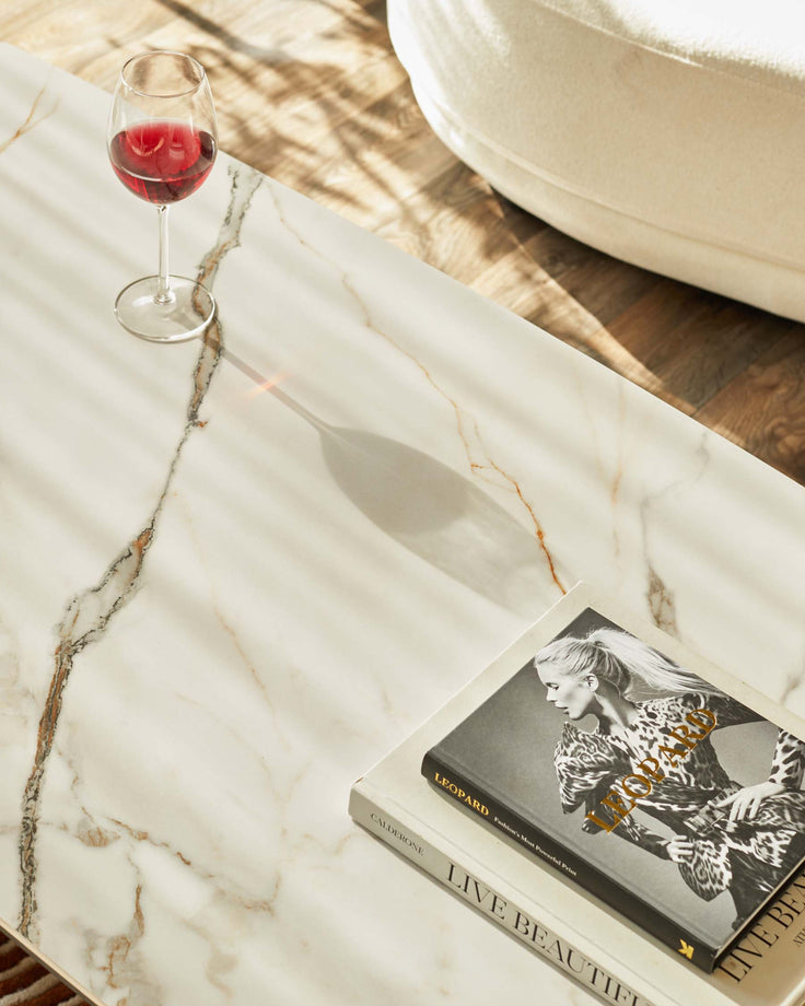 A marble coffee table with a glass of red wine and two books. There is a white pouffe in the background.