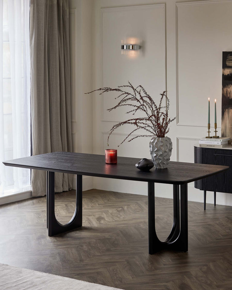 Elegant modern dining table with a dark wood finish and U-shaped legs, styled with decorative branches in a vase, set against a neutral-toned minimalist interior.