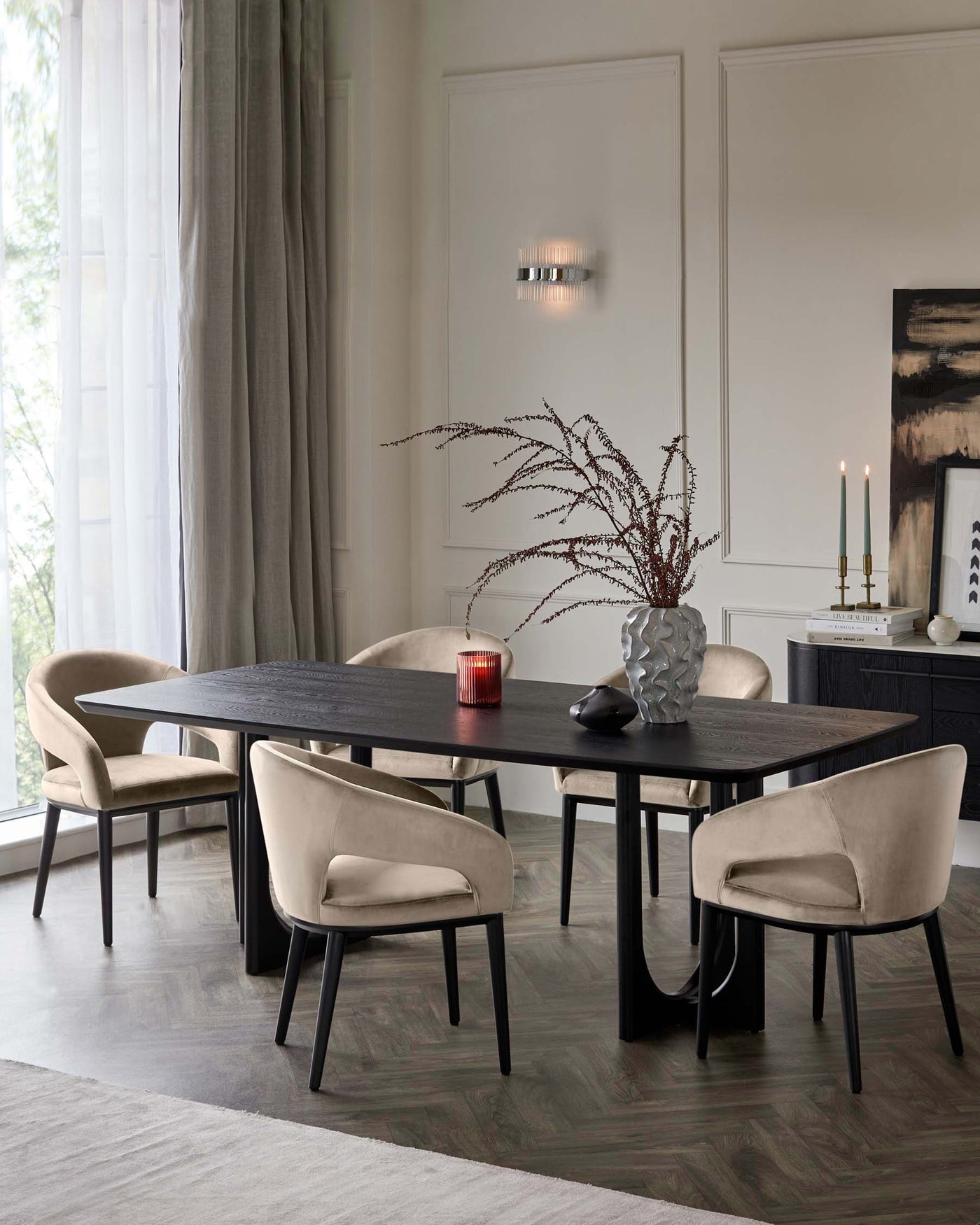 Elegant dining room with a modern, rectangular black wooden table and four plush beige upholstered chairs with dark wooden legs. The table is adorned with a decorative vase and candle holders. The setting is on a textured grey area rug over a herringbone-patterned floor.