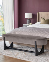A stylish taupe velvet bench with geometric dark wood legs sits at the foot of a bed in a modern bedroom.
