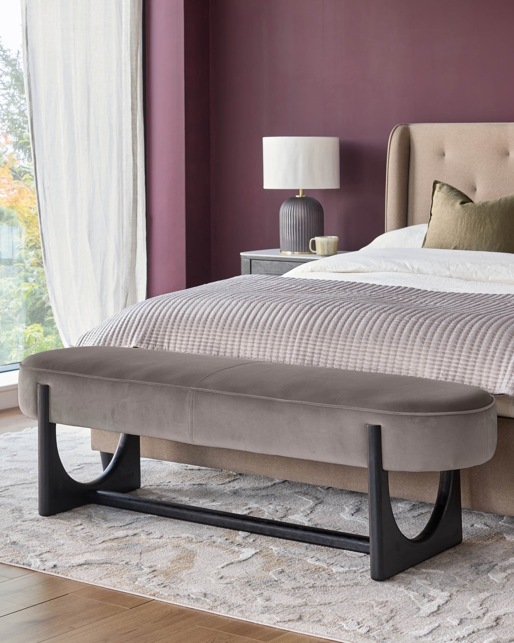 A stylish taupe velvet bench with geometric dark wood legs sits at the foot of a bed in a modern bedroom.