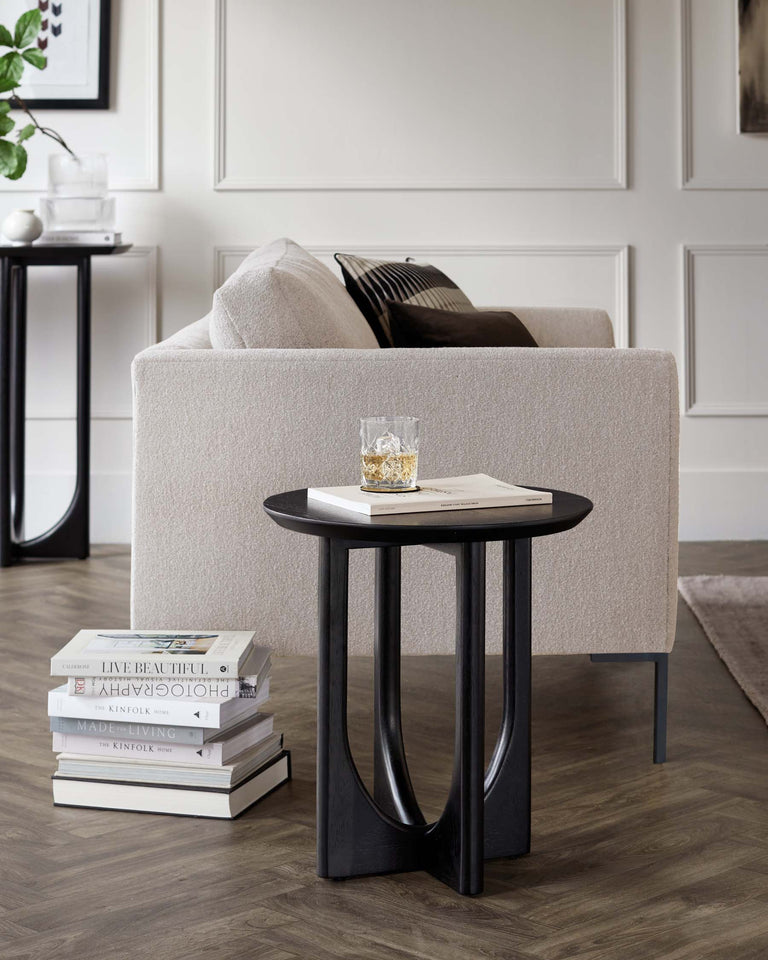 A modern round side table with a black finish and uniquely curved legs. Beside it, a partial view of a contemporary light beige sofa with plush cushions and a smooth fabric texture. A stack of decorative books is placed under the table, adding to the stylish home decor ambiance.