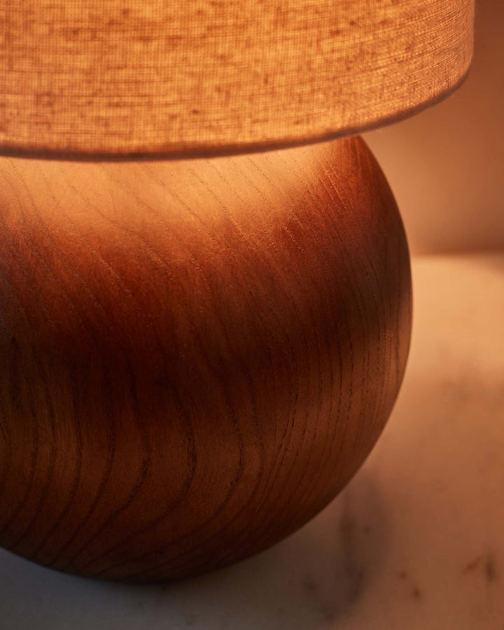 Round wooden lamp base with neutral lampshade sits on a marble-like surface. Warm ambient lighting.