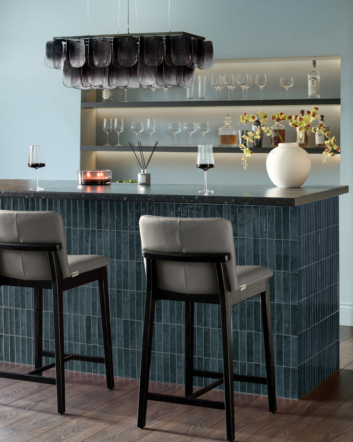 Otis bar stool in dark grey leather with black wooden frame, shown at modern bar counter with light grey upholstered backrest.