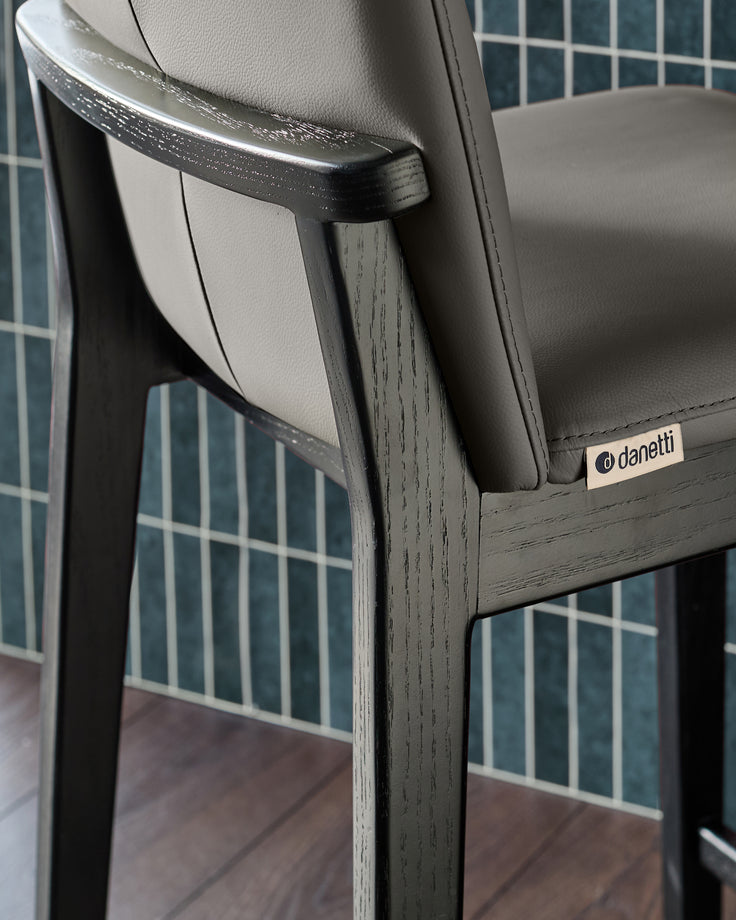 Dark grey wooden bar stool with grey leather seat and curved backrest, detail showing armrest and Danetti branding.
