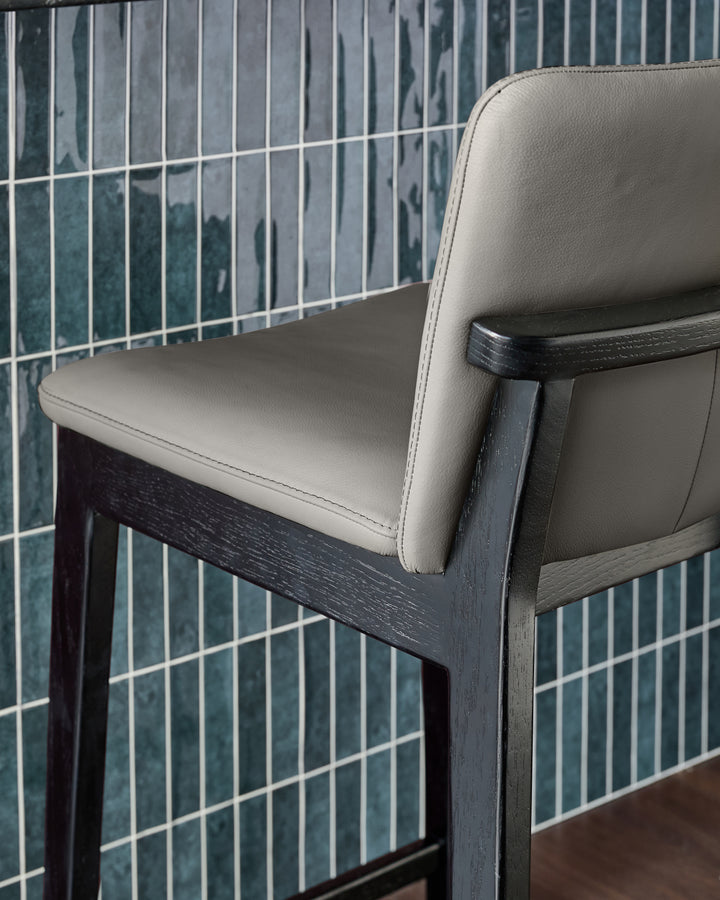 Otis bar stool in dark grey wood with cream leather seat and backrest, shown at an angle against tiled wall.