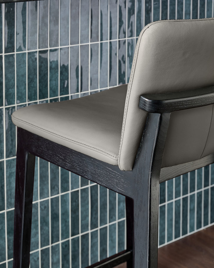 Otis bar stool in dark grey wood with cream leather seat and backrest, shown at an angle against tiled wall.