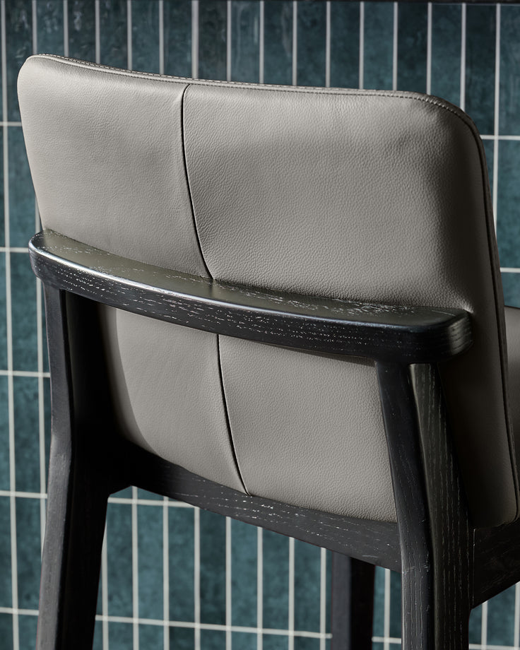 Otis bar stool in dark grey leather with black wooden frame, shown from side angle highlighting the backrest and seat.