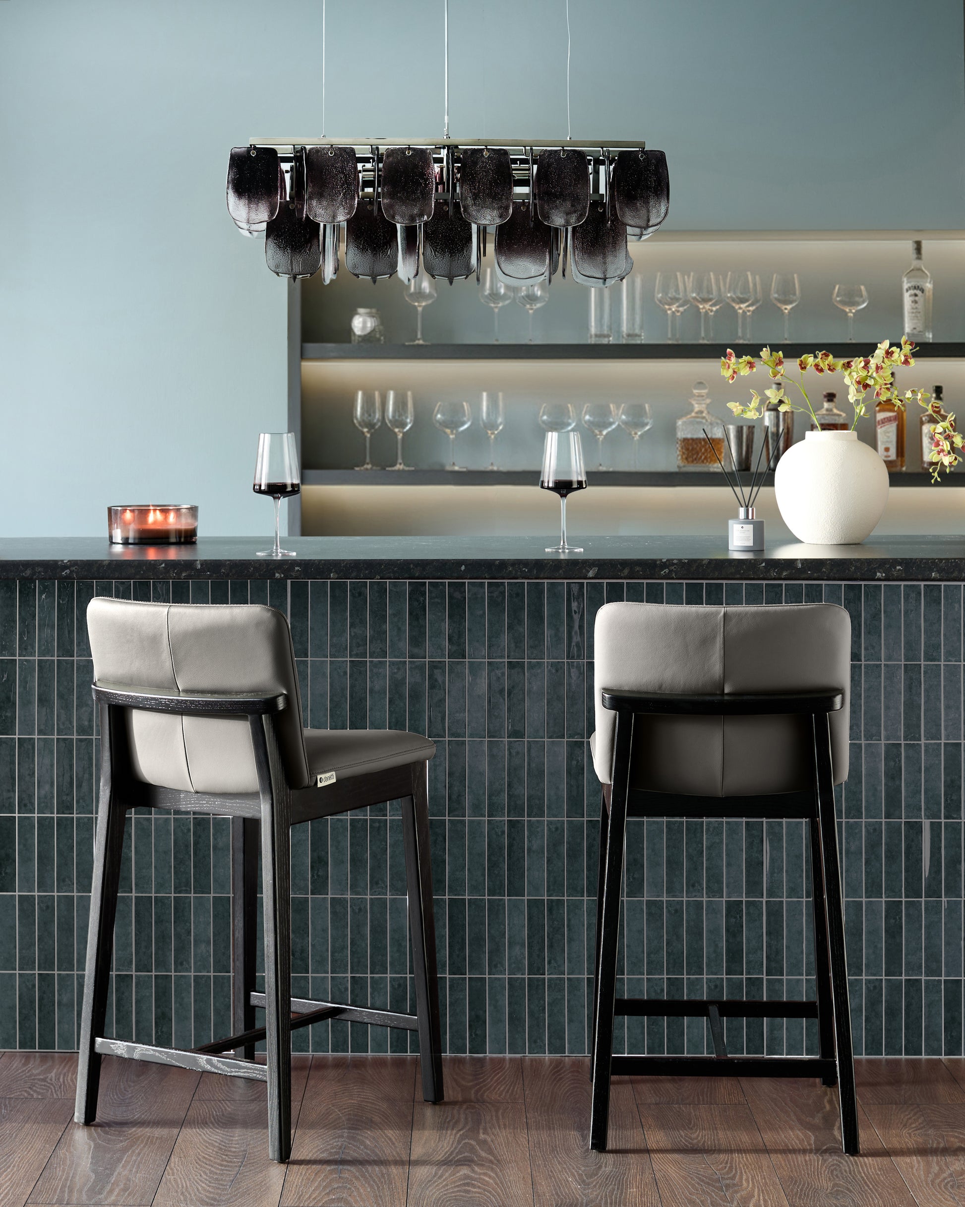 Otis bar stools in dark grey leather with black wooden frames, shown as a pair at a countertop in a modern bar setting.