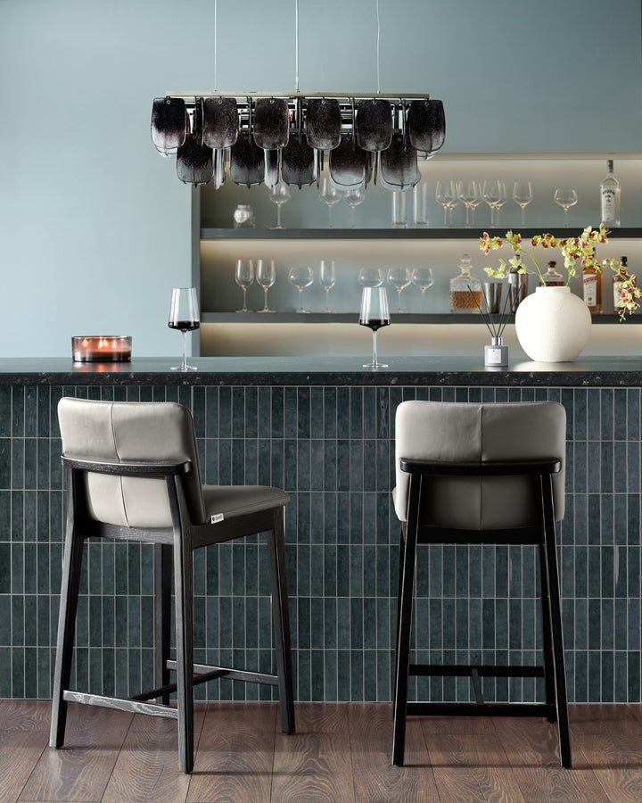 Otis bar stools in dark grey leather with black wooden frames, shown as a pair at a countertop in a modern bar setting.
