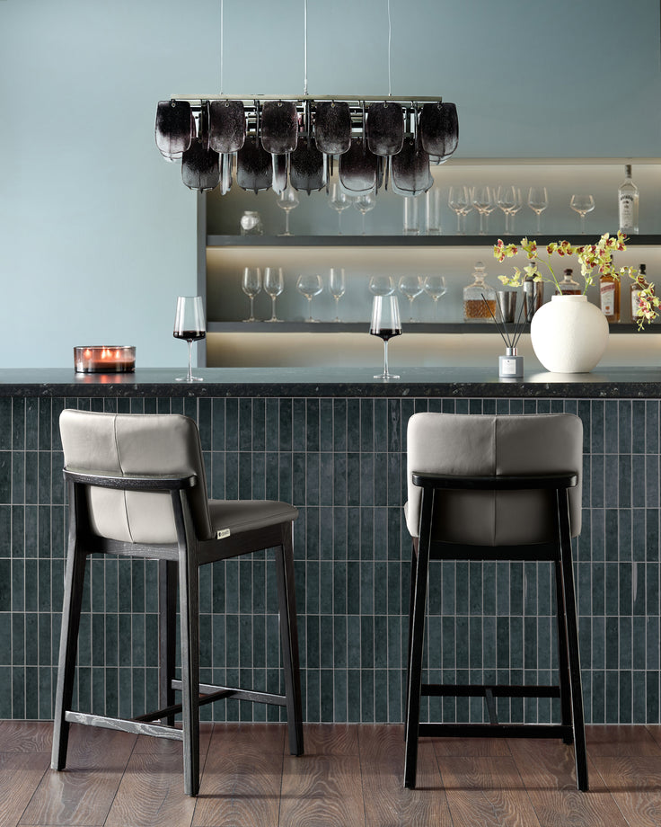 Otis bar stools in dark grey leather with black wooden frames, shown as a pair at a countertop in a modern bar setting.