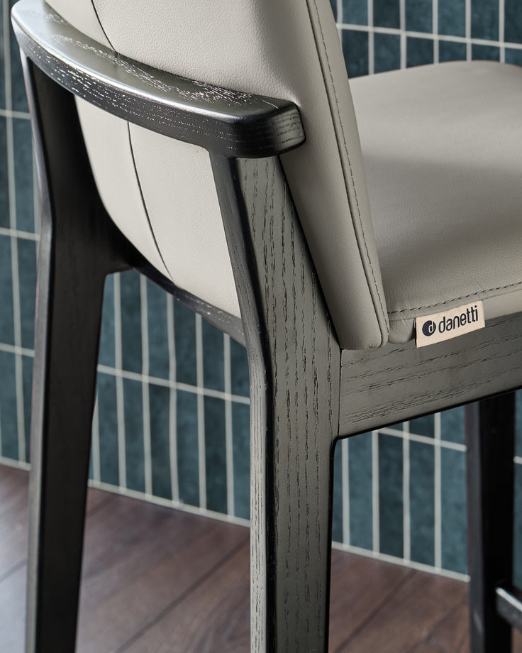 Otis bar stool with light grey leather seat, dark wood frame, curved backrest and armrest, close-up side view.