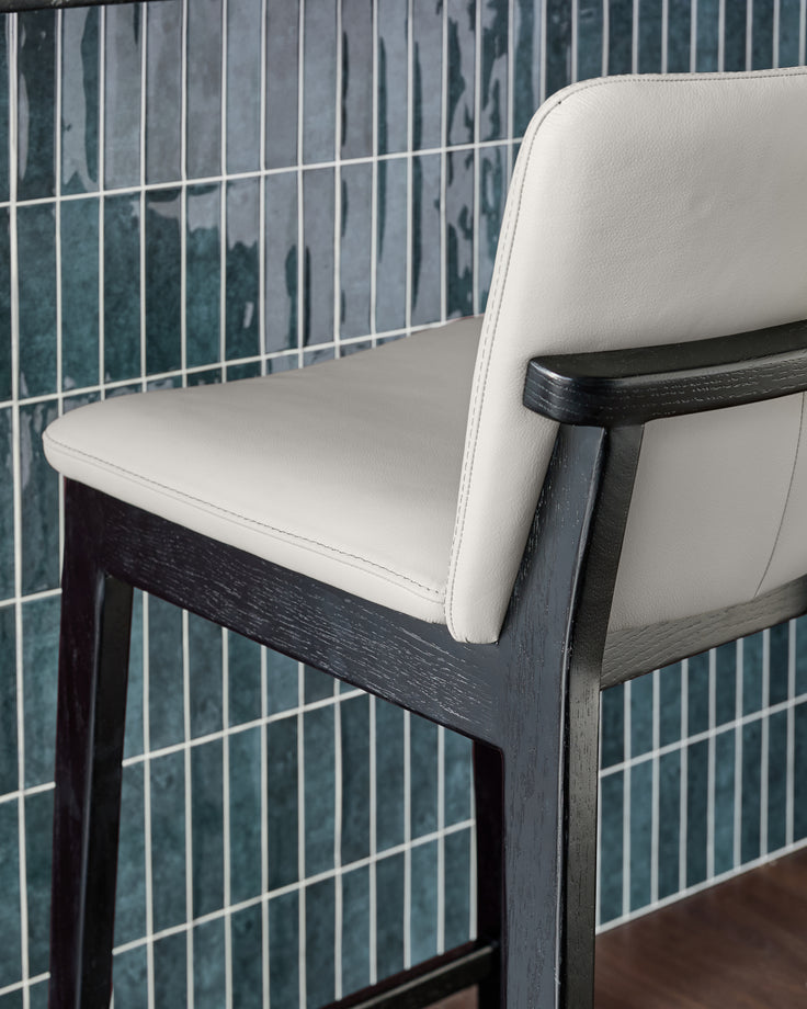 Otis bar stool with light grey leather seat and black wooden frame, shown at an angle against tiled backsplash.