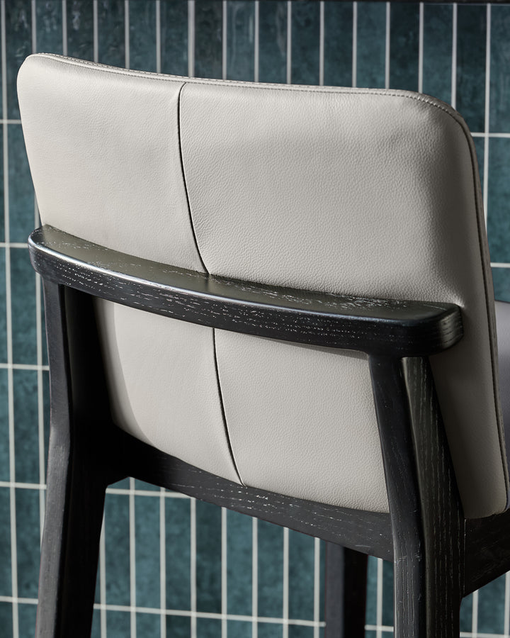 Otis bar stool with light grey leather seat and black wooden frame, shown from back angle against tiled wall.