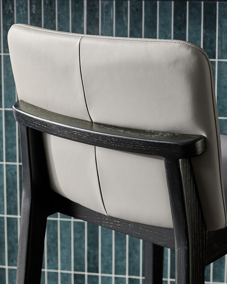 Otis bar stool with light grey leather seat and black wooden frame, shown from back angle against tiled wall.