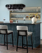 Otis bar stool in light grey leather with black wooden frame, shown at dark teal kitchen island counter.