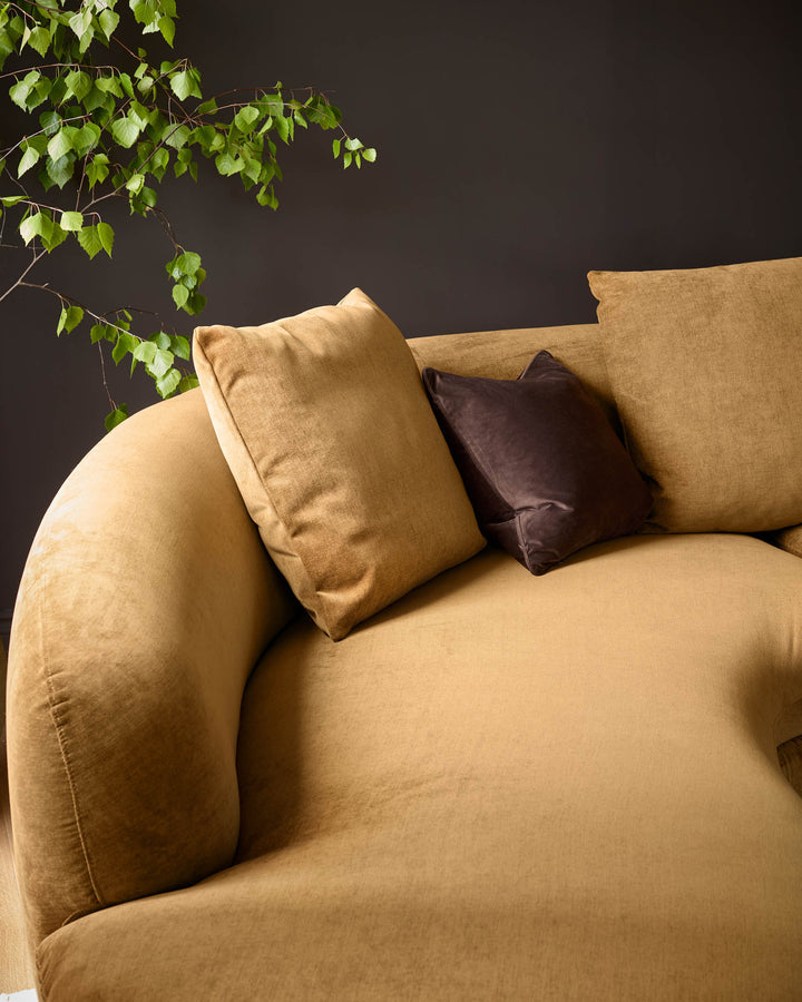 Ochre fabric left-hand corner chaise sofa with curved armrest, styled with tan and brown cushions against dark background with trailing ivy.