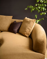 Ochre fabric right-hand corner chaise sofa with curved armrest, styled with tan and dark brown cushions against dark wall with trailing ivy.