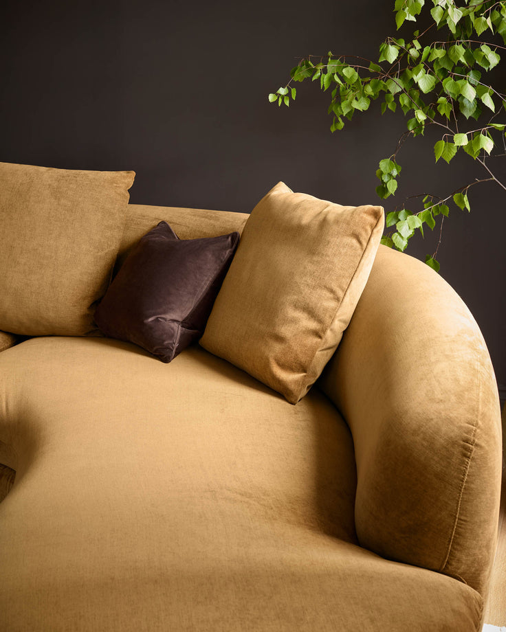 Ochre fabric right-hand corner chaise sofa with curved armrest, styled with tan and dark brown cushions against dark wall with trailing ivy.