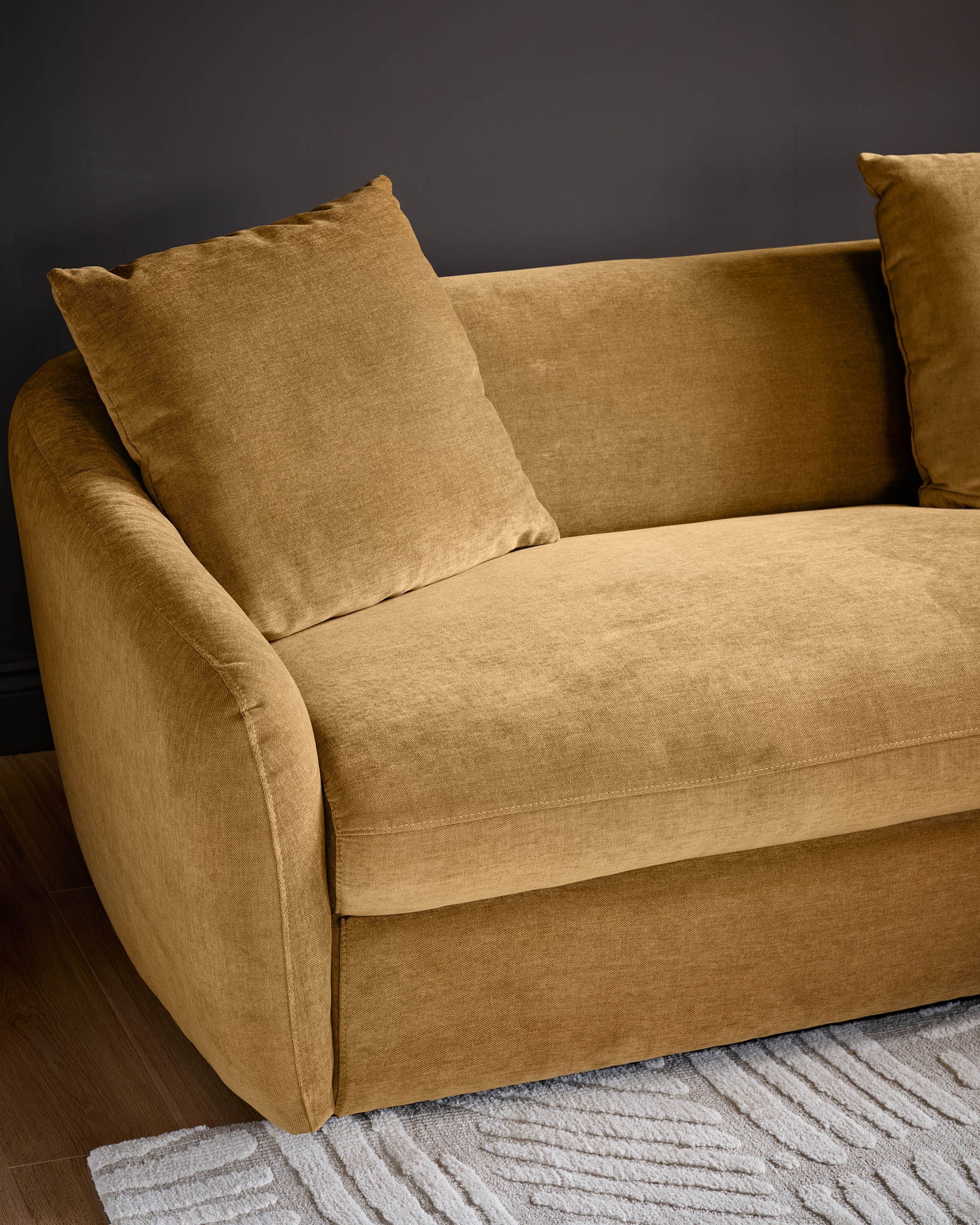 Ochre fabric right-hand corner chaise sofa with rounded arms and matching cushions, displayed against a dark background on a patterned rug.