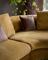 Ottilie right-hand corner chaise sofa in ochre fabric with brown and mauve decorative pillows, styled with trailing green foliage.