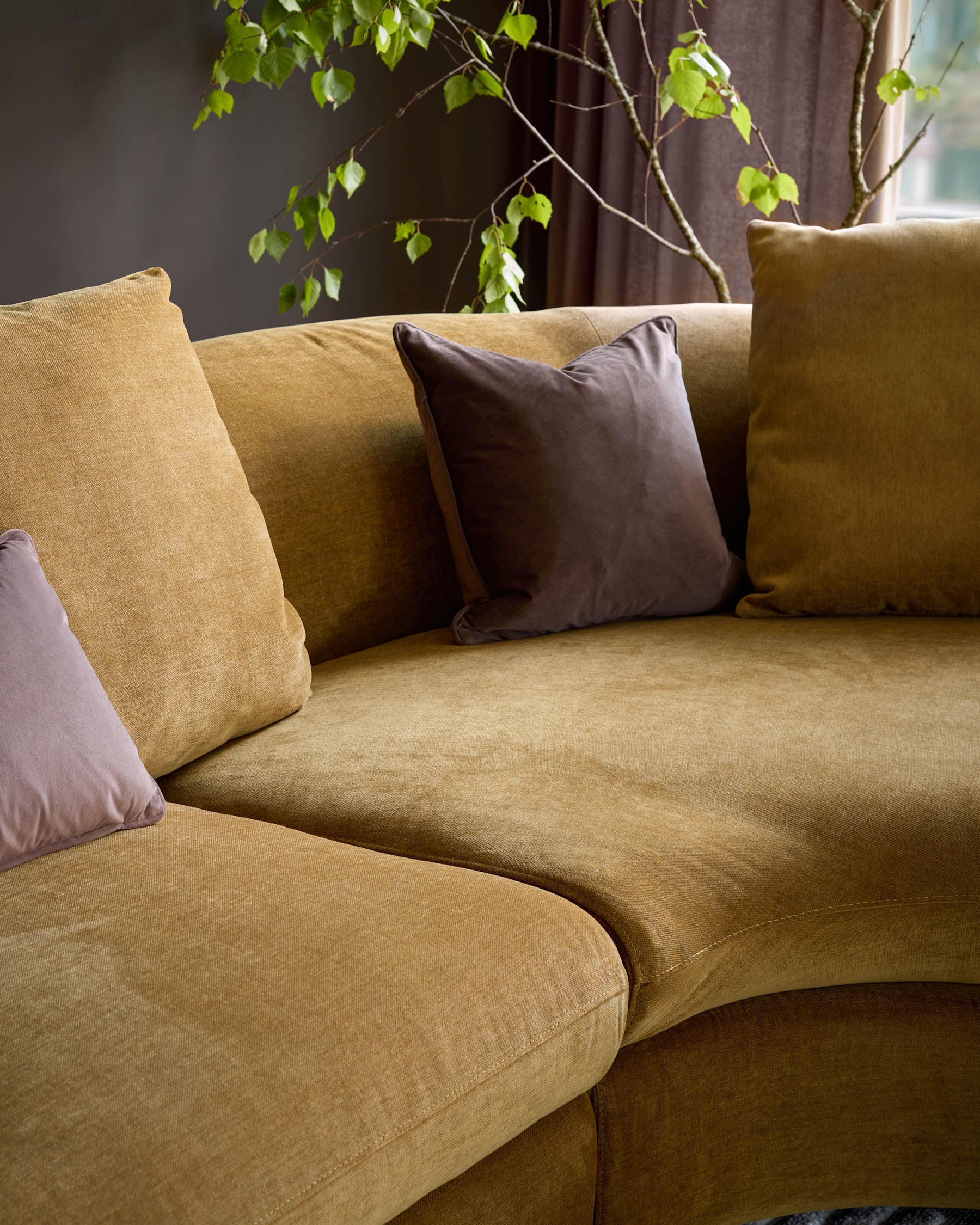 Ottilie right-hand corner chaise sofa in ochre fabric with brown and mauve decorative pillows, styled with trailing green foliage.