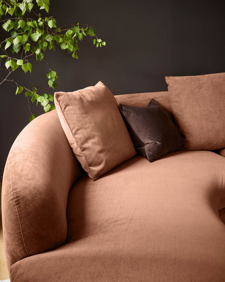 Ottilie left-hand corner chaise sofa in apricot fabric, angled view with brown scatter cushions and green foliage overhead.
