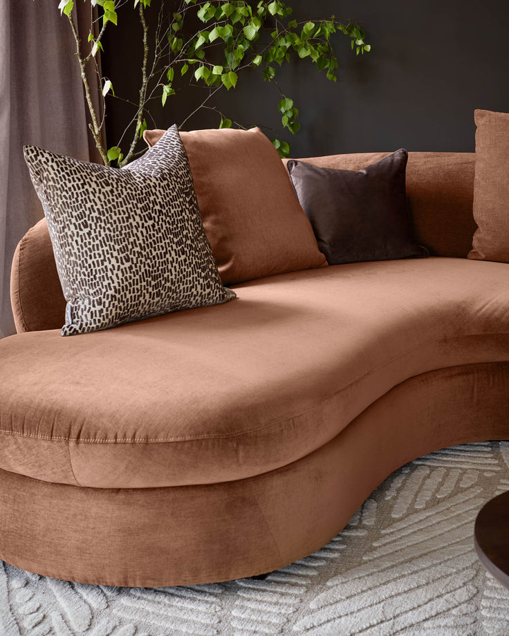 Apricot fabric left-hand corner chaise sofa with curved seating, styled with patterned and solid pillows, against dark wall with trailing plant.