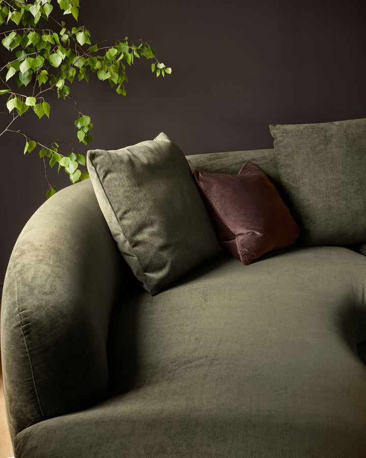 Olive green left-hand corner chaise sofa with curved armrests, styled with sage and burgundy cushions against a dark wall with trailing ivy.