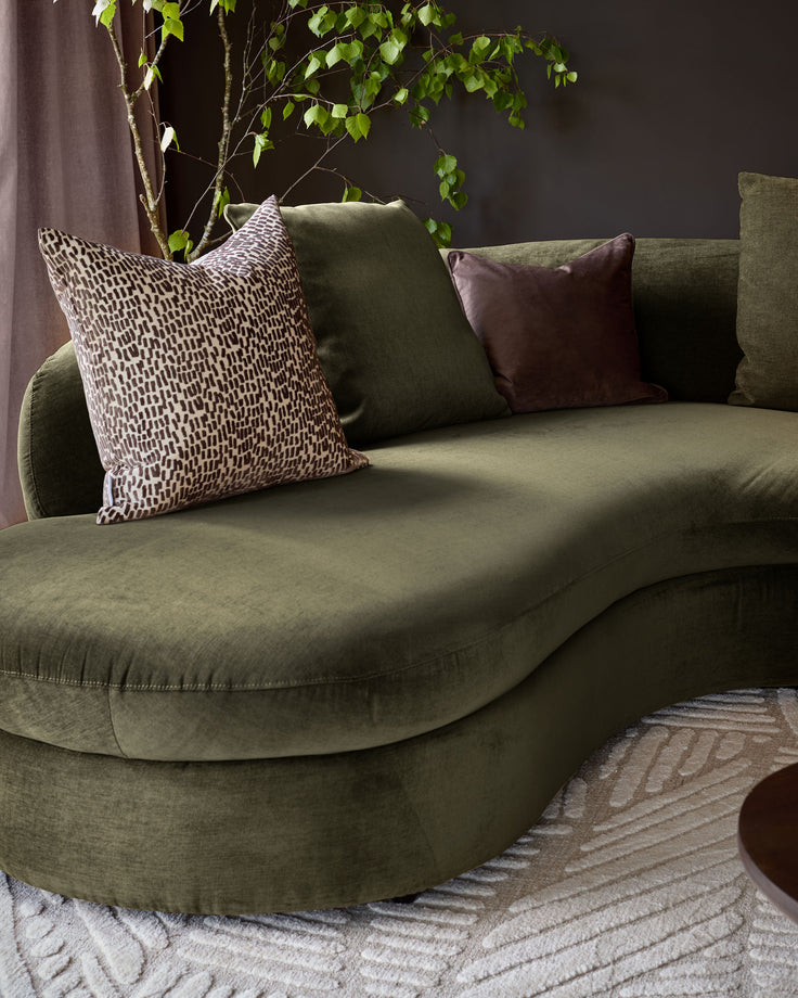 Ottilie left-hand corner chaise sofa in olive green fabric, styled with patterned and solid cushions, against dark wall with trailing plant.