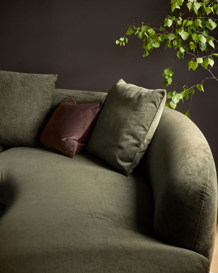 Olive green fabric right-hand corner chaise sofa with burgundy and sage cushions against dark wall with trailing ivy.
