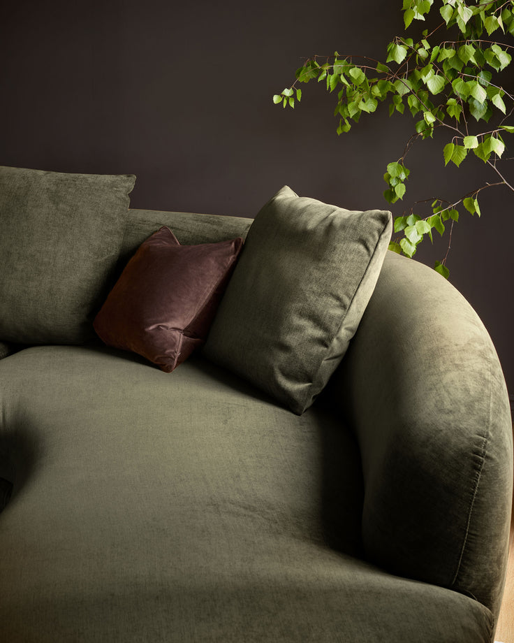 Olive green fabric right-hand corner chaise sofa with burgundy and sage cushions against dark wall with trailing ivy.