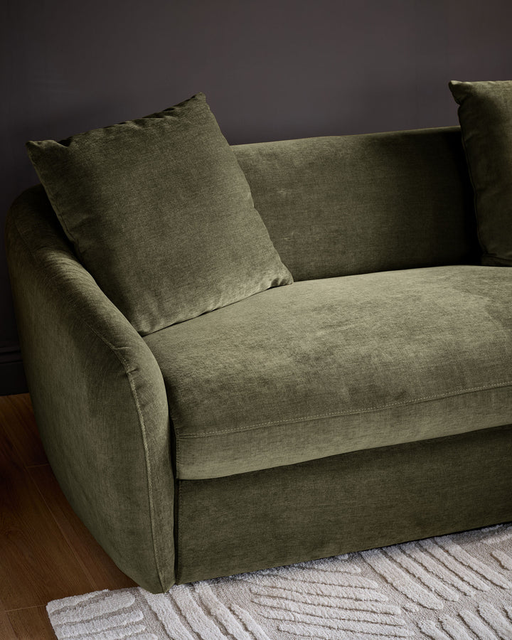 Olive green fabric right-hand corner chaise sofa with curved arms and matching throw pillows against a dark wall.