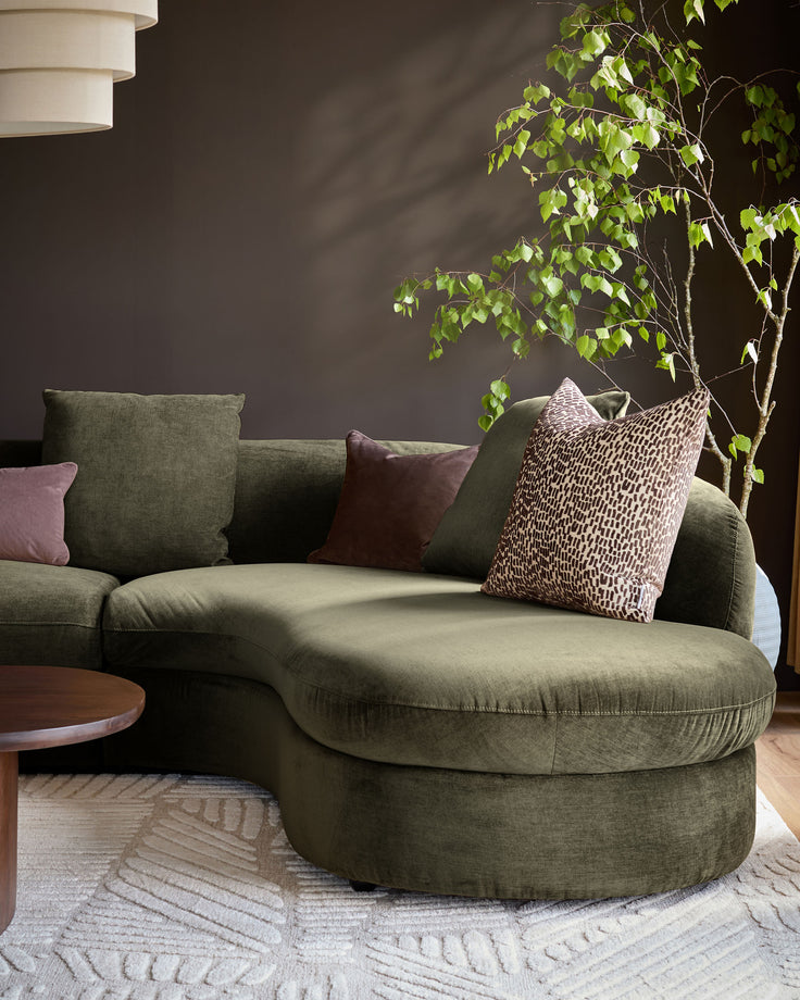 Olive-green fabric corner chaise sofa with curved rounded armrests, styled with decorative pillows in a modern living room setting.