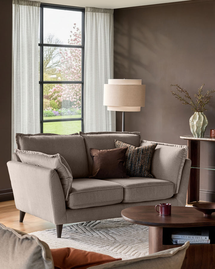Otto 2-seater sofa in mocha fabric displayed in a modern living room with throw pillows and wooden legs.