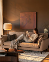 Otto 3-seater sofa in mocha fabric, displayed in a warm living room setting with a woman reading on it.
