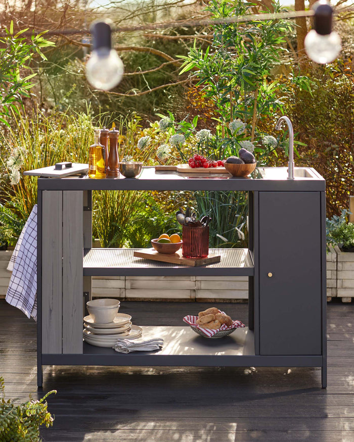 Outdoor kitchen island with modern design, featuring a built-in sink with a high arc faucet, a dark grey countertop, and charcoal-coloured storage cabinets. The lower shelf displays dishware and textiles. Set in a garden with ambient lighting.