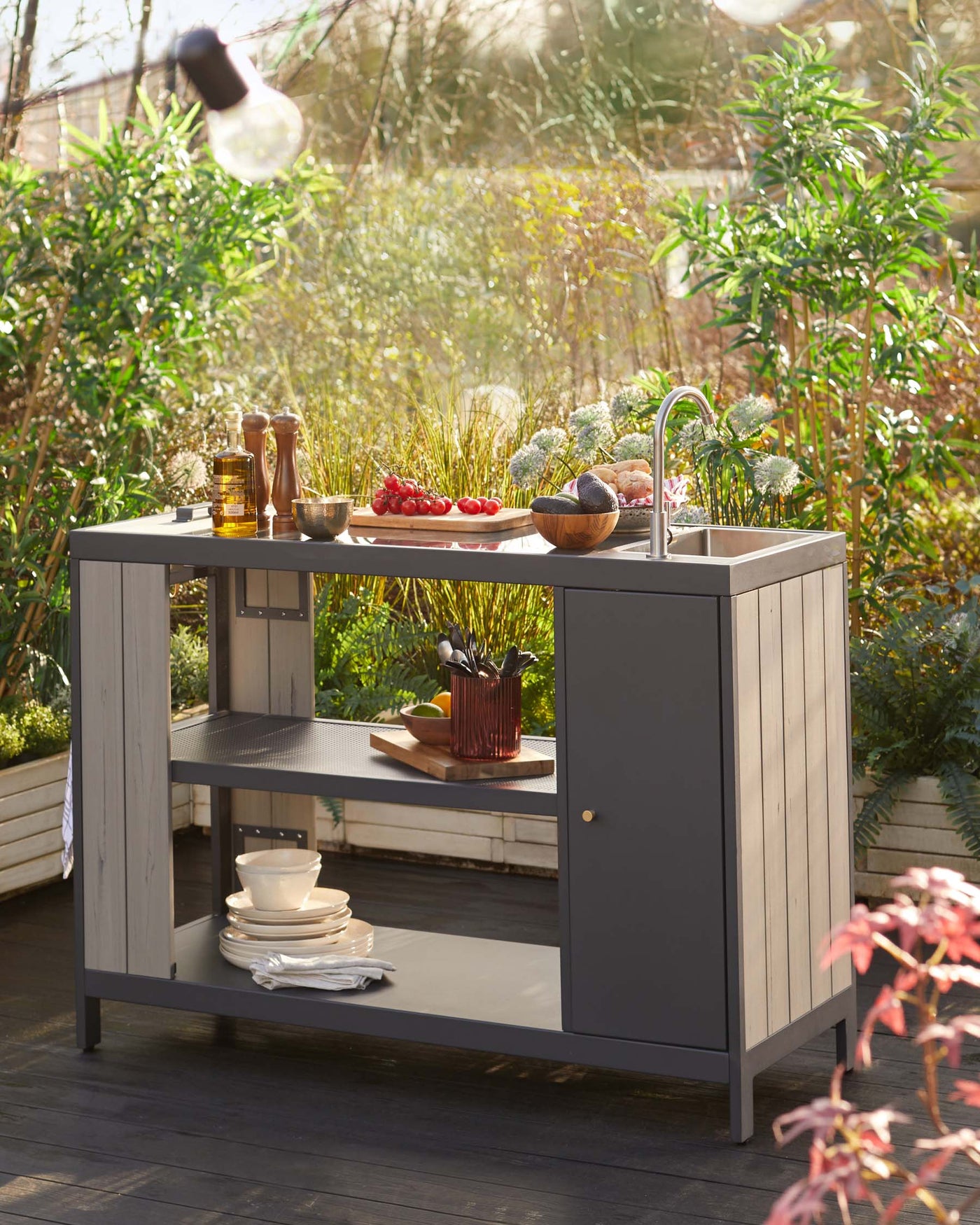 Outdoor cooking station featuring a modern, charcoal-grey finish with a built-in sink, an open lower shelf, and a side storage cabinet, displayed in a garden setting.