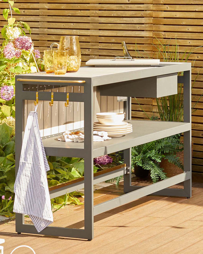Outdoor garden potting table in grey with two shelves, featuring gardening tools, hanging utensils, a striped towel, and arranged with tableware, a pitcher, and drinking glasses atop its work surface, set against a lush garden and wood slat backdrop.