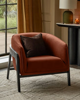 Palmer occasional chair in toffee-red fabric with dark wooden frame and black leather accent pillow, shown in styled interior setting.