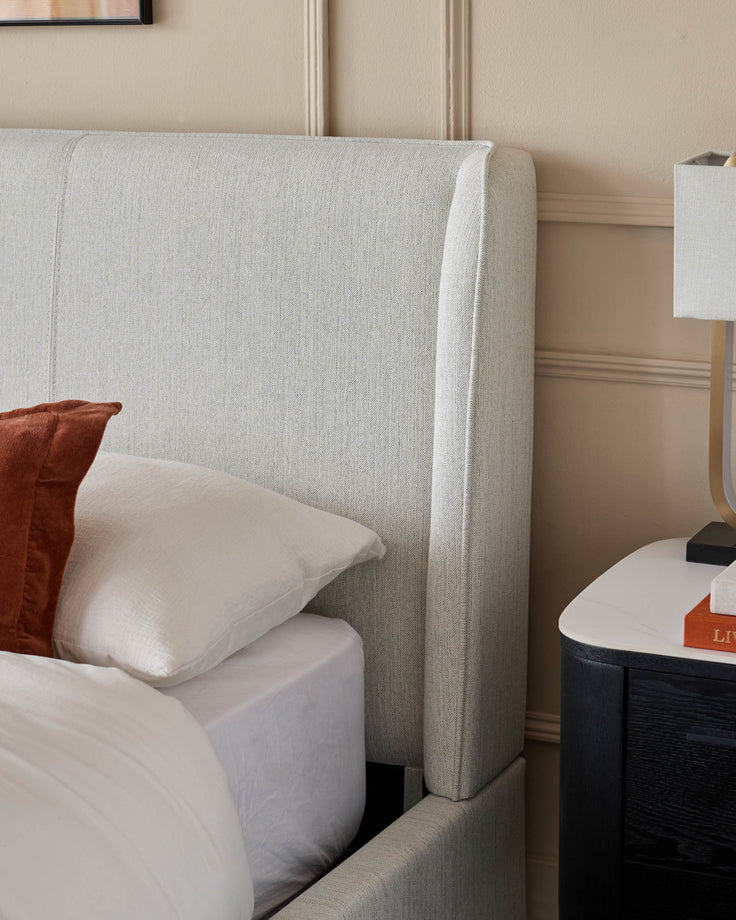 A grey upholstered bed with pillows and a dark wood nightstand with a marble top.