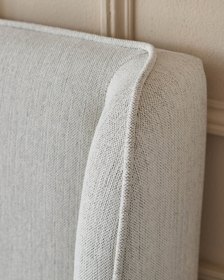 Close up of an upholstered headboard, featuring light grey woven fabric with a subtle texture.