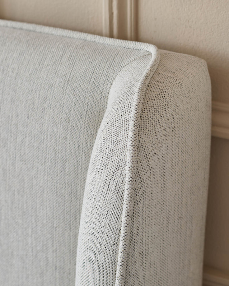 Close-up of a textured light grey upholstered headboard with piped edge detailing against a neutral wall.