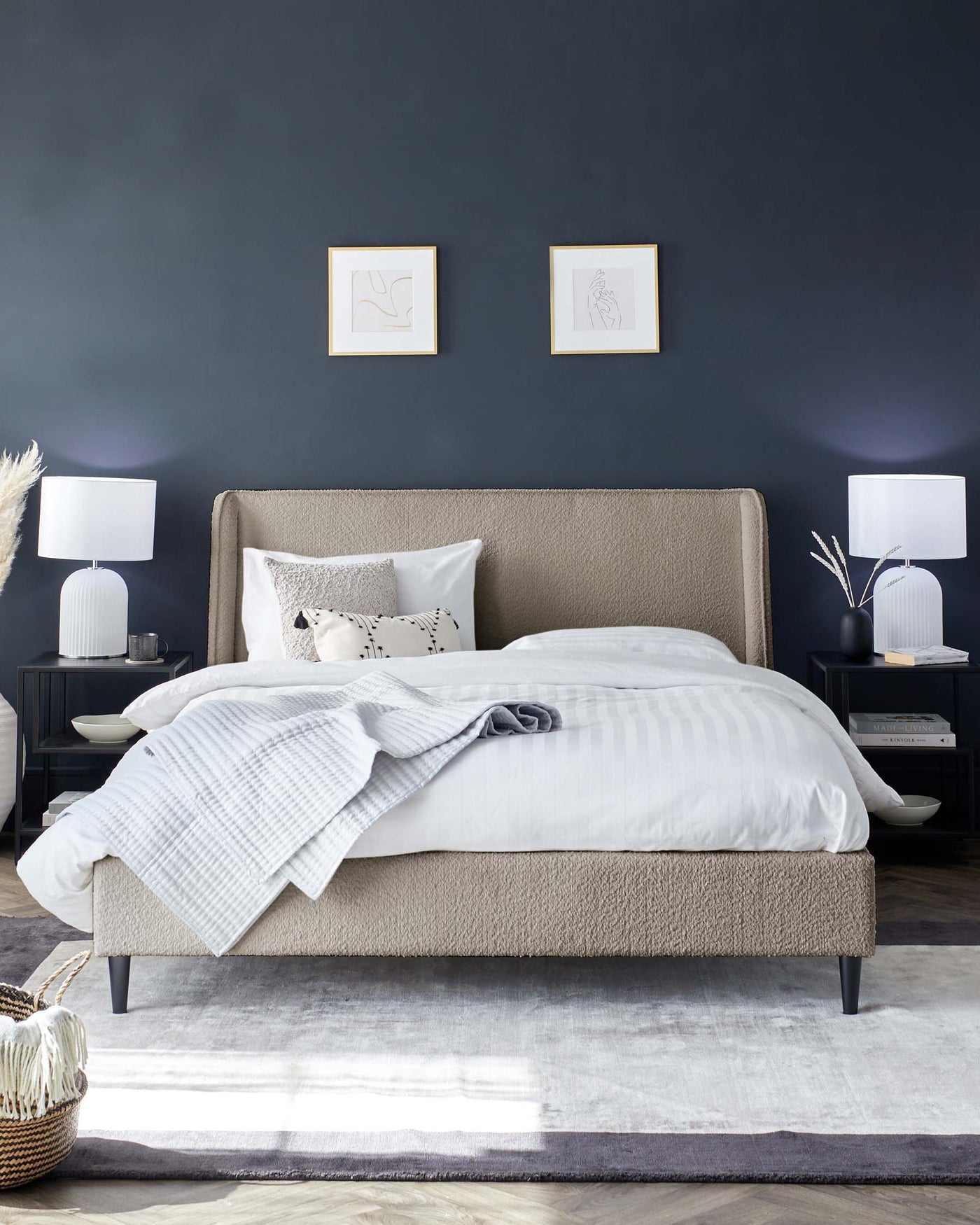 Modern bedroom featuring an upholstered platform bed in a neutral beige fabric with a high, cushioned headboard and dark tapered legs. Coordinating bedside tables in black with elegant white table lamps, and a soft grey floor rug complete the contemporary aesthetic.