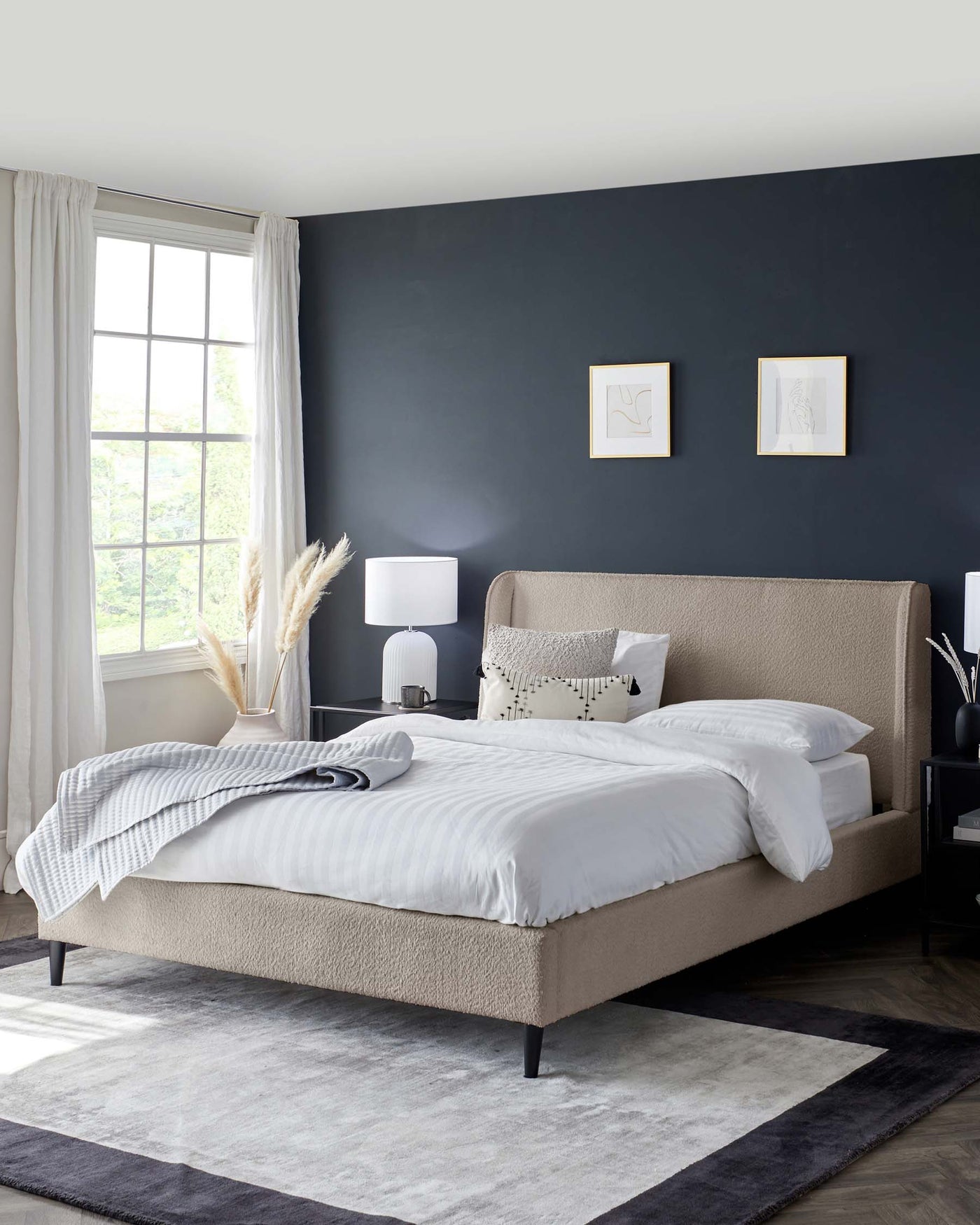 Contemporary upholstered queen-sized bed with a high headboard in a neutral beige tone, white bedding with a striped throw blanket, on a two-tone grey area rug. Matching bedside tables with black bases and a white table lamp on one side.